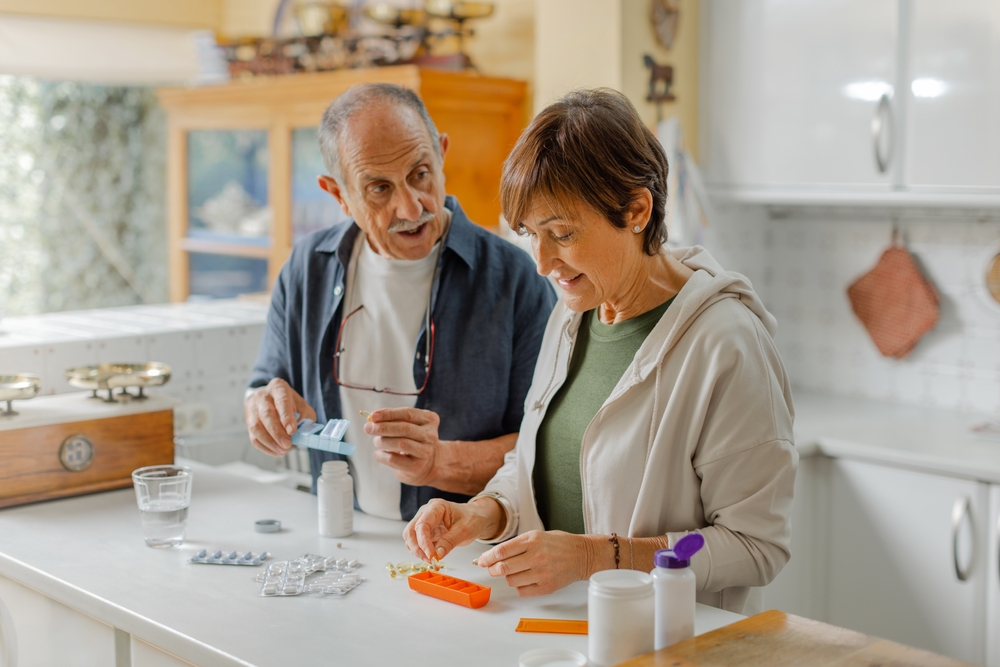 A man and a woman stand at their kitchen counter. Each of them are measuring out pills and putting them into pill organizers.